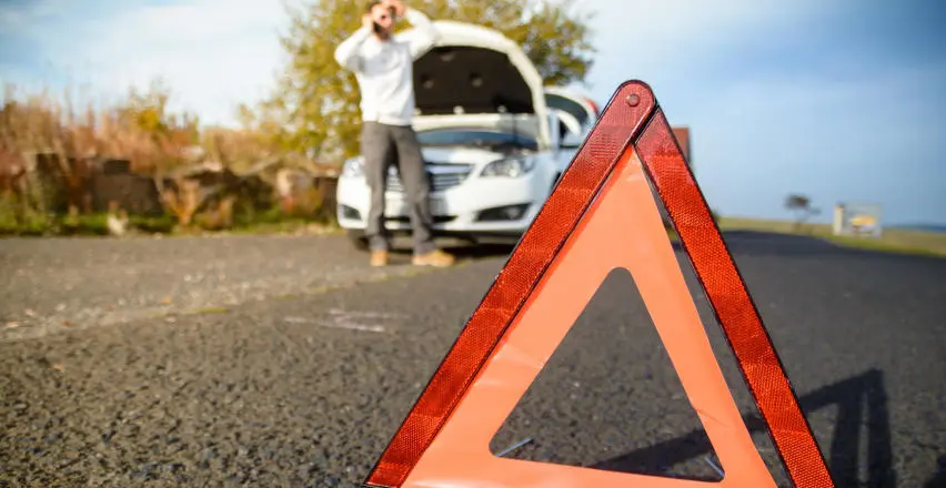 obraz przedstawiający rozłożony trójkąt ostrzegawczy oraz mężczyzne w tle trzymającego się za głowę, dzwoniącego przy otawrtej masce auta